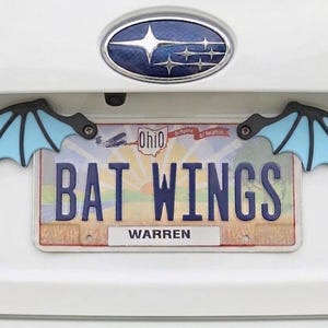 May include: A white car's rear with a license plate that reads "BAT WINGS" and bat wing accents. The bat wings are black and light blue. The license plate is from Ohio and has the name "WARREN" on it.