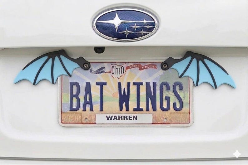 May include: A white car's rear with a license plate that reads "BAT WINGS" and bat wing accents. The bat wings are black and light blue. The license plate is from Ohio and has the name "WARREN" on it.