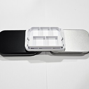 May include: Three rectangular tin boxes, one black, one silver, and one white with a grid of six compartments. The white box is open and the other two are closed.