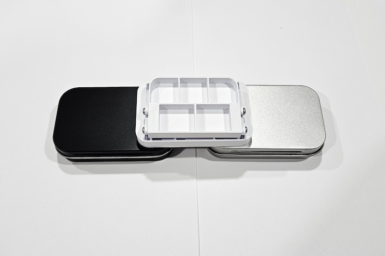 May include: Three rectangular tin boxes, one black, one silver, and one white with a grid of six compartments. The white box is open and the other two are closed.