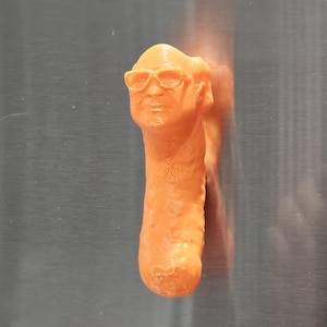 May include: Orange 3D-printed figurine of a man's head wearing glasses, atop a long, cylindrical body. The figurine is a solid orange color and appears to be made of plastic. The background is a plain, metallic surface.