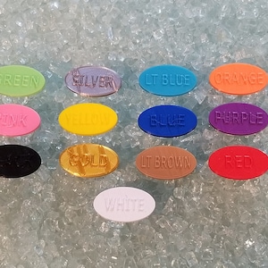 May include: A set of 14 oval-shaped plastic counters in various colours, including green, silver, light blue, orange, pink, yellow, blue, purple, black, gold, light brown, red, and white. Each counter has a colour name printed on it.