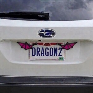 May include: A white Subaru Outback vehicle featuring a custom license plate frame with two stylized, pointed pink and black bat-like wings attached to the top corners. The Ohio license plate displays the text DRAGON2 in bold blue capital letters.