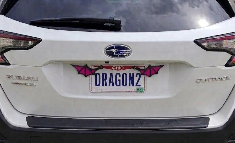May include: A white Subaru Outback vehicle featuring a custom license plate frame with two stylized, pointed pink and black bat-like wings attached to the top corners. The Ohio license plate displays the text DRAGON2 in bold blue capital letters.