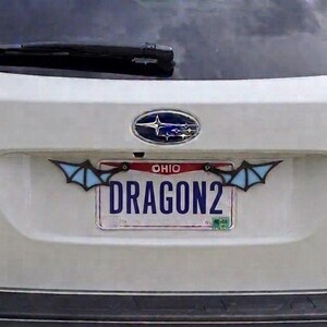 May include: A white Subaru Outback vehicle featuring a rear license plate from Ohio with the text DRAGON2. The license plate is framed by two decorative light blue and black wing-shaped attachments mounted on the top corners of the plate frame.
