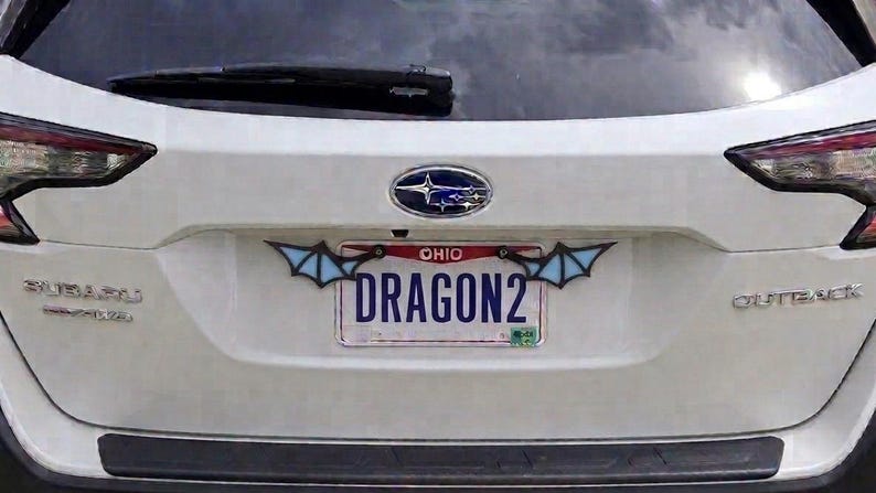 May include: A white Subaru Outback vehicle featuring a rear license plate from Ohio with the text DRAGON2. The license plate is framed by two decorative light blue and black wing-shaped attachments mounted on the top corners of the plate frame.