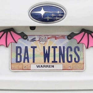 May include: Pink and black bat wing car accessories attached to a license plate that reads "BAT WINGS" in blue letters. The license plate is from Ohio and has the name "WARREN" at the bottom.