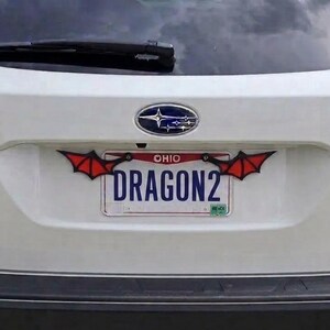 May include: A white Subaru Outback rear hatch featuring an Ohio license plate with the text DRAGON2. The license plate frame is decorated with two black and red bat-like wings attached to the top corners, extending outward from the plate.