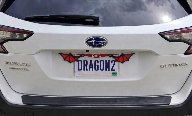 May include: A white Subaru Outback rear hatch featuring an Ohio license plate with the text DRAGON2. The license plate frame is decorated with two black and red bat-like wings attached to the top corners, extending outward from the plate.