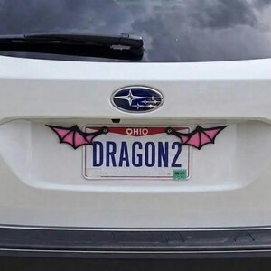 May include: A white Subaru Outback vehicle featuring a custom license plate frame with black and pink bat wing accents attached to the top corners. The Ohio license plate displays the text DRAGON2 in blue capital letters against a white background.