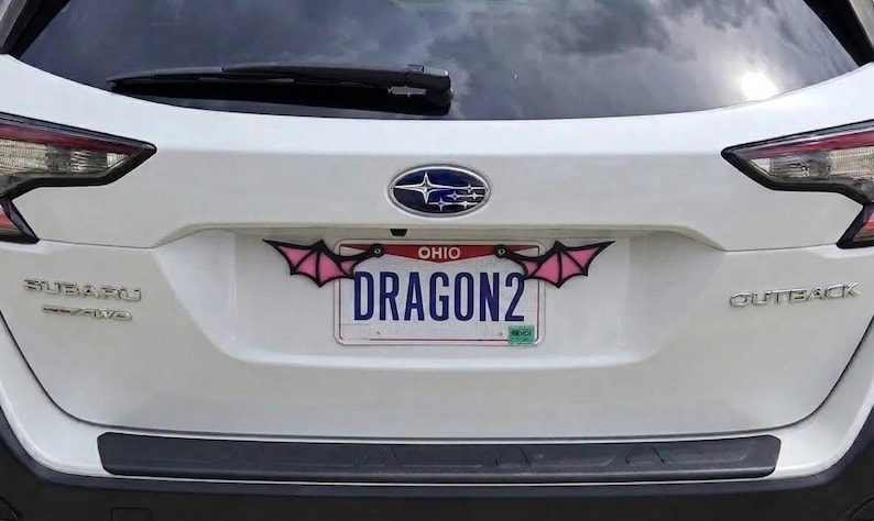 May include: A white Subaru Outback vehicle featuring a custom license plate frame with black and pink bat wing accents attached to the top corners. The Ohio license plate displays the text DRAGON2 in blue capital letters against a white background.