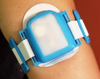 May include: A blue and white medical device with a clear plastic window. The device is worn on the arm and has a strap with a buckle.