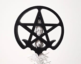 May include: A black glittery circular pentagram ornament featuring a small bat silhouette at the bottom center. The center of the pentagram is filled with a bundle of thin, white, shredded paper-like material that extends below the frame.