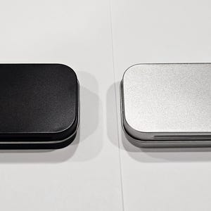 May include: Two small rectangular metal tins, one black and one silver, with a slightly rounded top and a hinged lid.