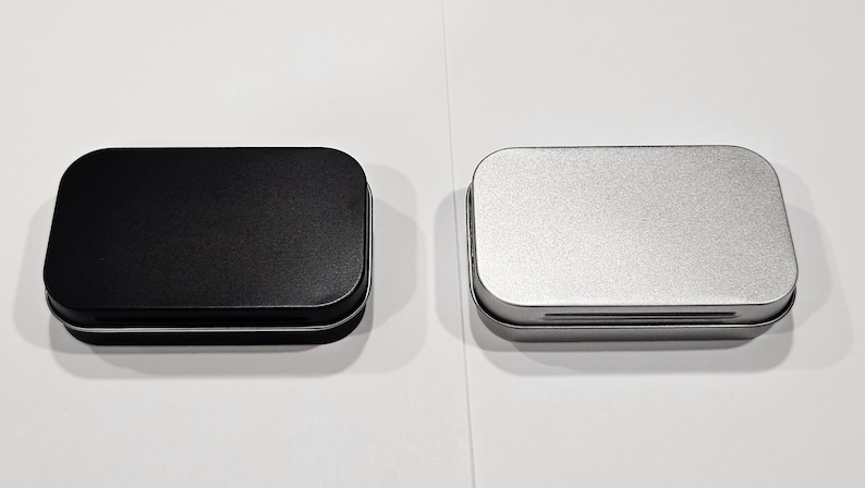 May include: Two small rectangular metal tins, one black and one silver, with a slightly rounded top and a hinged lid.