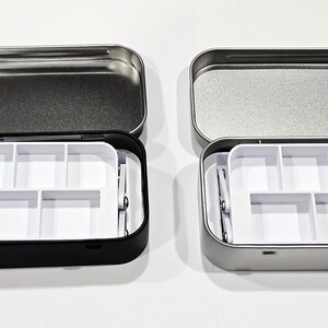 May include: Two small metal tins with lids, one black and one silver, each containing a white plastic tray with six compartments. The trays are designed to hold small items like beads, buttons, or other craft supplies.