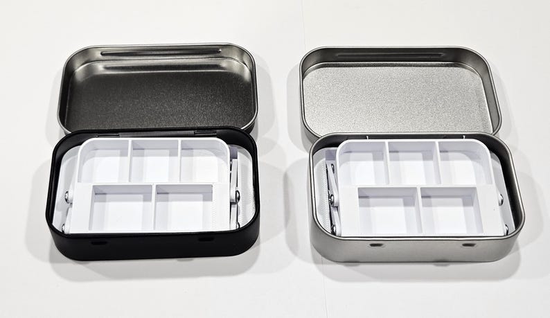 May include: Two small metal tins with lids, one black and one silver, each containing a white plastic tray with six compartments. The trays are designed to hold small items like beads, buttons, or other craft supplies.