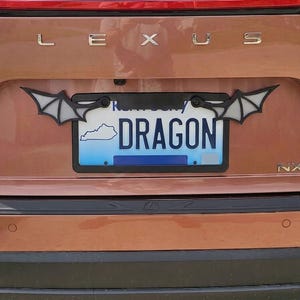 May include: A black license plate frame featuring stylized bat wings extending from the top corners. The frame is mounted on a copper-colored Lexus NX350 vehicle, surrounding a Kentucky license plate with the text DRAGON and a state outline.