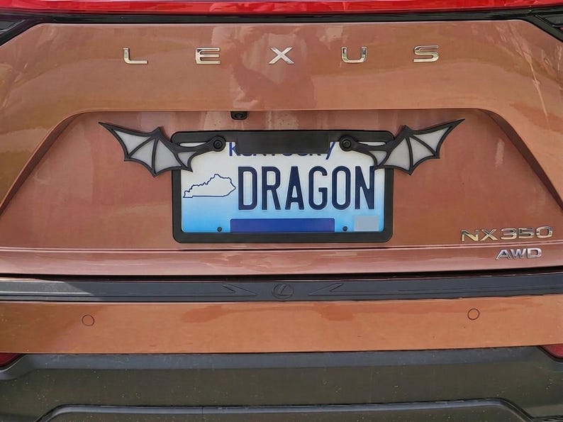 May include: A black license plate frame featuring stylized bat wings extending from the top corners. The frame is mounted on a copper-colored Lexus NX350 vehicle, surrounding a Kentucky license plate with the text DRAGON and a state outline.