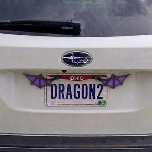 May include: A white Subaru Outback vehicle featuring a license plate frame with purple and black bat wing accents attached to the top corners. The Ohio license plate displays the text DRAGON2 in bold blue letters against a white background.