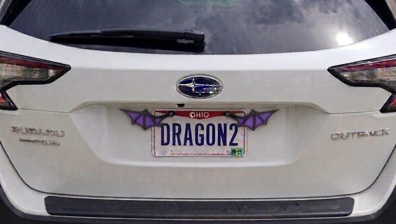 May include: A white Subaru Outback vehicle featuring a license plate frame with purple and black bat wing accents attached to the top corners. The Ohio license plate displays the text DRAGON2 in bold blue letters against a white background.