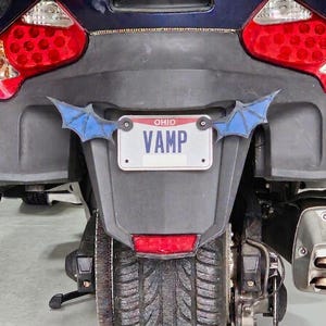 May include: The rear view of a dark blue motorcycle featuring a white Ohio license plate with the text VAMP in blue letters. The plate is flanked by two decorative blue bat wing accents. The motorcycle has large red taillights and a dual exhaust pipe.