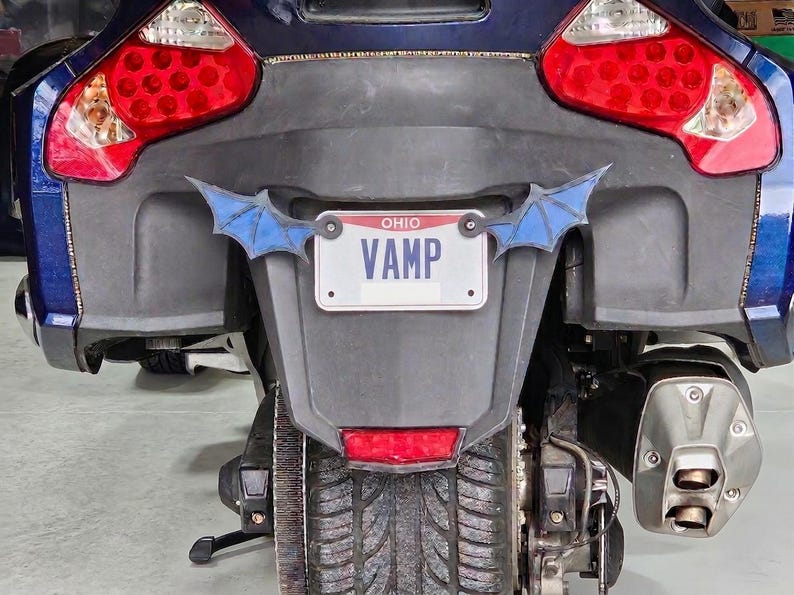 May include: The rear view of a dark blue motorcycle featuring a white Ohio license plate with the text VAMP in blue letters. The plate is flanked by two decorative blue bat wing accents. The motorcycle has large red taillights and a dual exhaust pipe.