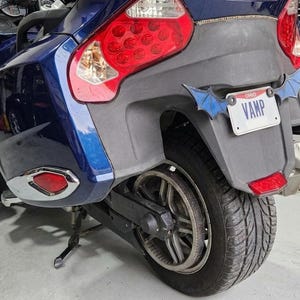May include: A dark blue motorcycle rear section featuring a black fender, a large red taillight, and a license plate that reads VAMP with blue bat-wing shaped accents attached to the sides of the plate frame, mounted on a black tire and wheel.