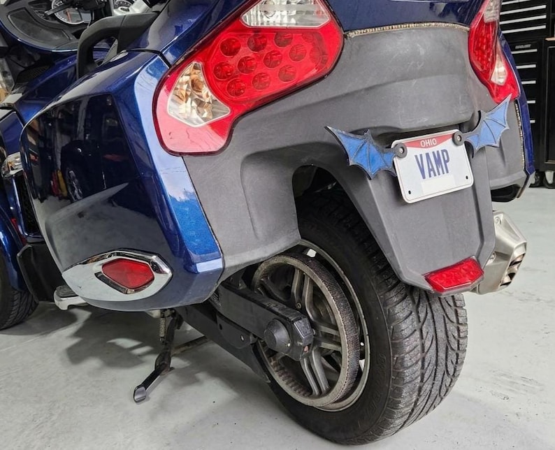 May include: A dark blue motorcycle rear section featuring a black fender, a large red taillight, and a license plate that reads VAMP with blue bat-wing shaped accents attached to the sides of the plate frame, mounted on a black tire and wheel.