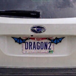 May include: A white Subaru Outback vehicle featuring a custom license plate frame with black and blue bat-like wings attached to the top corners. The license plate displays the text OHIO at the top and the word DRAGON2 in large blue letters.