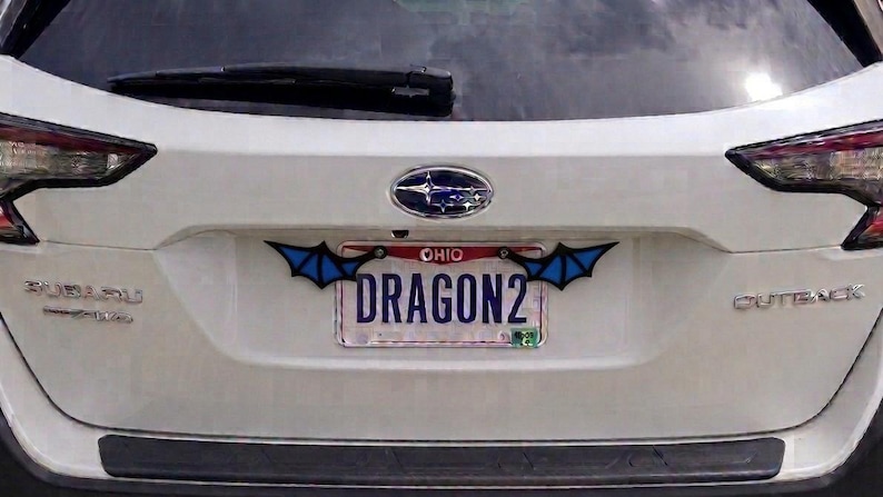 May include: A white Subaru Outback vehicle featuring a custom license plate frame with black and blue bat-like wings attached to the top corners. The license plate displays the text OHIO at the top and the word DRAGON2 in large blue letters.