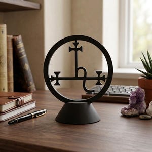 May include: A matte black circular sculpture featuring an intricate alchemical symbol of Saturn centered within the ring, mounted on a solid conical base. The design includes a vertical cross and a horizontal bar with cross-tipped ends.