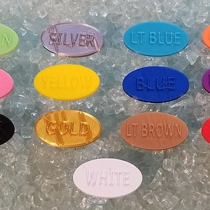 May include: A collection of eleven oval-shaped plastic tags in various colors including green, silver, light blue, orange, pink, yellow, blue, purple, black, gold, light brown, red, and white, each embossed with its corresponding color name.