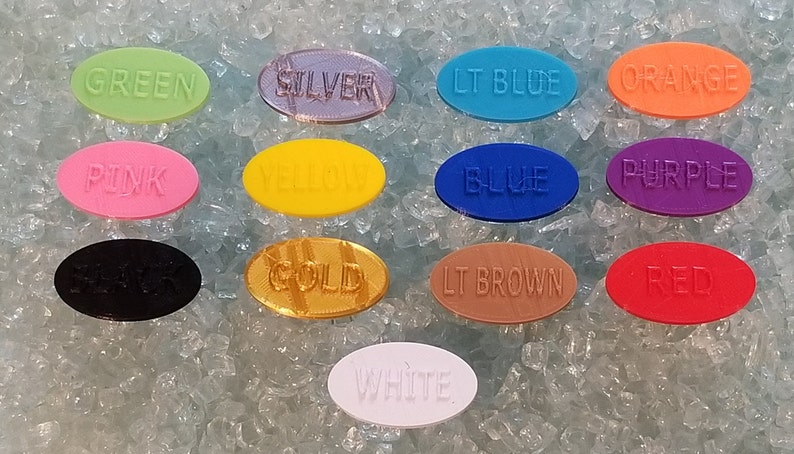 May include: A collection of eleven oval-shaped plastic tags in various colors including green, silver, light blue, orange, pink, yellow, blue, purple, black, gold, light brown, red, and white, each embossed with its corresponding color name.