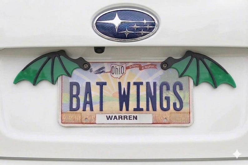 May include: A white car's trunk with a license plate that reads "BAT WINGS" and two green and black bat wing decorations. The license plate is from Ohio and has the name "WARREN" at the bottom.