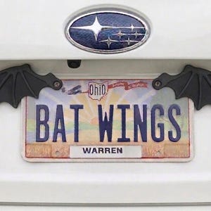 May include: Black bat wing license plate frame. The license plate reads "BAT WINGS" with the city of Warren at the bottom. The wings are attached to the car with screws.