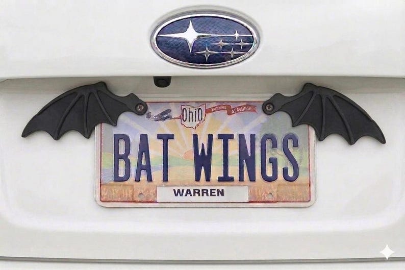 May include: Black bat wing license plate frame. The license plate reads "BAT WINGS" with the city of Warren at the bottom. The wings are attached to the car with screws.