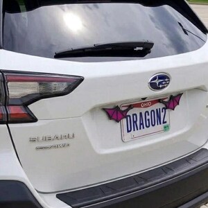 May include: A white Subaru vehicle features a license plate frame adorned with two purple and black bat-like wing attachments. The license plate displays the text OHIO and DRAGON2 in blue lettering against a white background with a small green sticker.