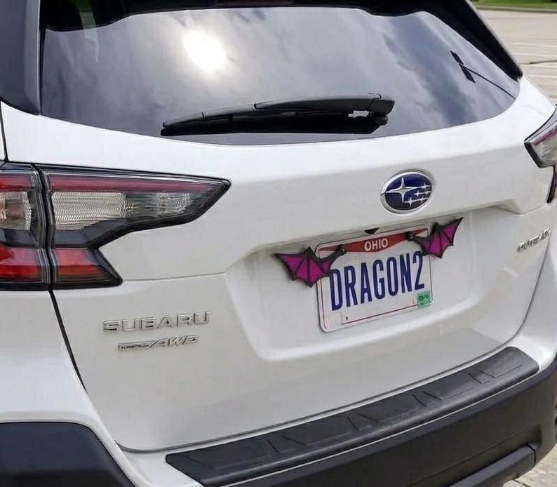 May include: A white Subaru vehicle features a license plate frame adorned with two purple and black bat-like wing attachments. The license plate displays the text OHIO and DRAGON2 in blue lettering against a white background with a small green sticker.