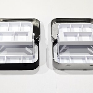 May include: Two metal storage boxes with a white plastic insert. The box on the left is black and the box on the right is silver.