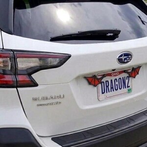 May include: The rear view of a white Subaru vehicle featuring a license plate frame decorated with red and black bat-like wings. The Ohio license plate displays the text DRAGON2 in blue lettering, positioned below the Subaru logo and rear wiper.