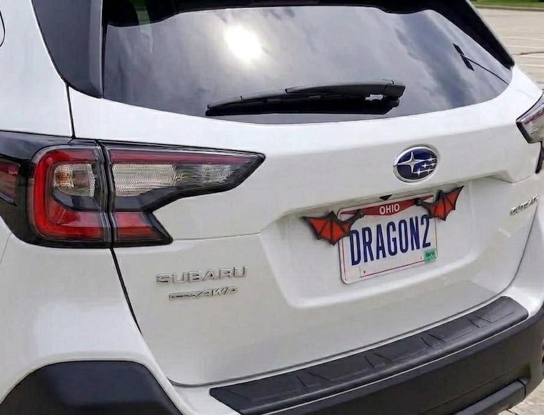 May include: The rear view of a white Subaru vehicle featuring a license plate frame decorated with red and black bat-like wings. The Ohio license plate displays the text DRAGON2 in blue lettering, positioned below the Subaru logo and rear wiper.