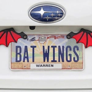 May include: Red and black bat wing license plate frame attached to a white car. The license plate reads "BAT WINGS" with the city of Warren displayed below. The Ohio license plate is visible.