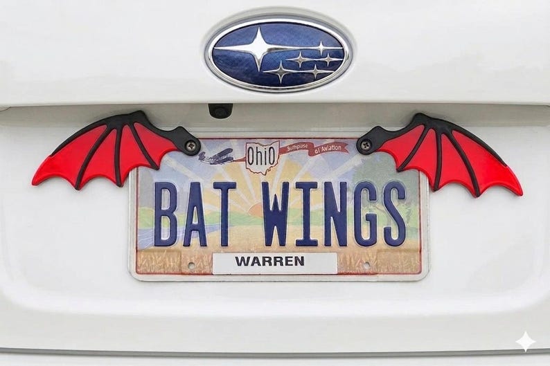 May include: Red and black bat wing license plate frame attached to a white car. The license plate reads "BAT WINGS" with the city of Warren displayed below. The Ohio license plate is visible.