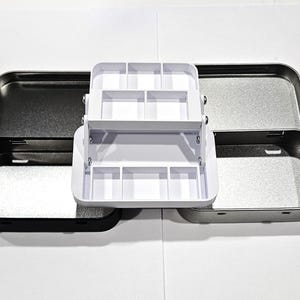 May include: A white plastic organizer with compartments for small items, sitting inside two metal tins. The tins are silver and black.