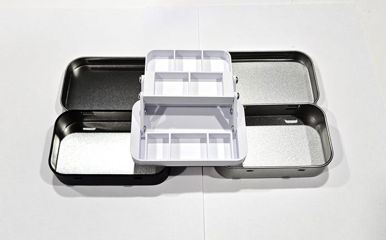 May include: A white plastic organiser with compartments for small items, sitting inside two metal tins. The tins are silver and black.