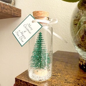 Könnte beinhalten: Eine kleine Glasflasche mit Korken, die einen Miniatur-Weihnachtsbaum und weißes, schneegleiches Material enthält. Ein Etikett mit "Merry Christmas & a Happy New Year" ist mit einem weißen Band befestigt. Die Flasche steht auf einer Holzoberfläche.