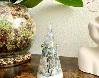 May include: A white cone-shaped ring holder with a clear resin top filled with dried flowers and glitter. A silver ring with clear stones sits on top of the holder.