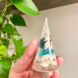 Op de afbeelding: Een transparante, kegelvormige papieren gewicht van hars met een ontwerp van blauwe en witte schelpen. De papieren gewicht is gevuld met kleine witte schelpen, gouden glitter en een blauwe glazen kraal.