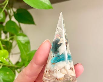 May include: A clear cone-shaped resin paperweight with a blue and white seashell design. The paperweight is filled with small white seashells, gold glitter, and a blue glass bead.